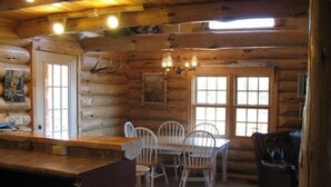 A log cabin interior with a wooden dining table, white chairs, a dark leather couch, and a chandelier. - Welcome to 'The Log Cab-Inn' - Where Great Memories Are Made (Atlantic Mine)