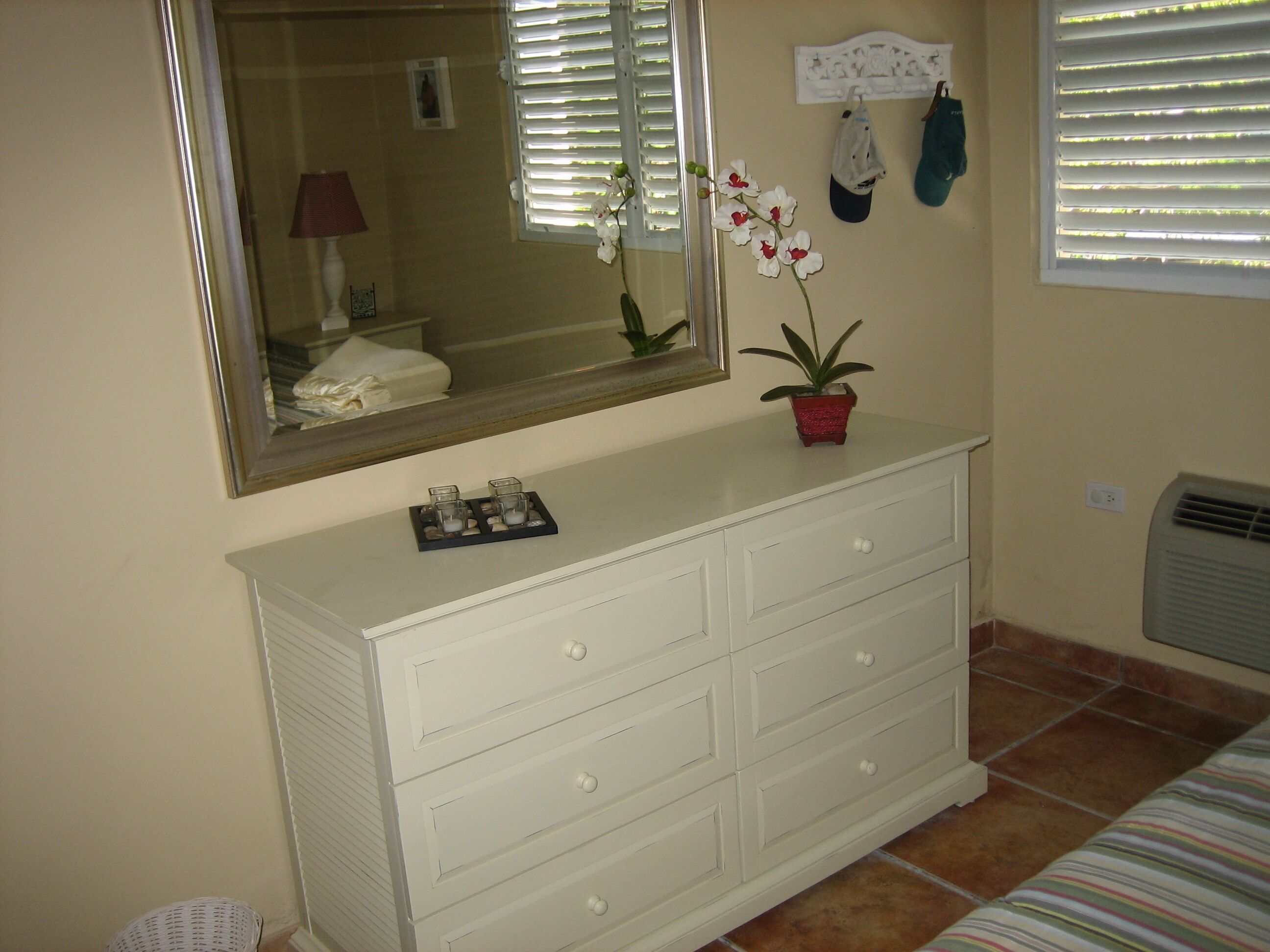 Une commode blanche avec des tiroirs, un miroir au-dessus, une petite orchidée en pot et une fenêtre avec des stores.