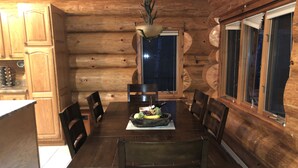 A wooden cabin interior with a dining table set for four, surrounded by chairs. A fruit bowl is placed on the table, and there is a window with curtains on the right side. - Custom Log Lake House on Wynne Lake  with Pontoon Boat. Summer 1 week Sat to Sat (Aurora)