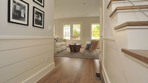A hallway with a staircase, framed black and white photos on the wall, a round coffee table, and a chair. - Nantucket Art House - In Town ~ Heated Pool ~ Includes Guest House!  (Nantucket)