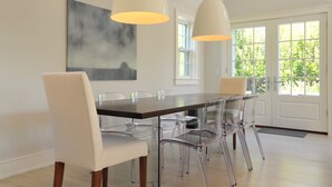 A dining area with a wooden table and clear chairs, pendant lights, a large abstract painting, and a view of greenery through the door. - Nantucket Art House - In Town ~ Heated Pool ~ Includes Guest House!  (Nantucket)
