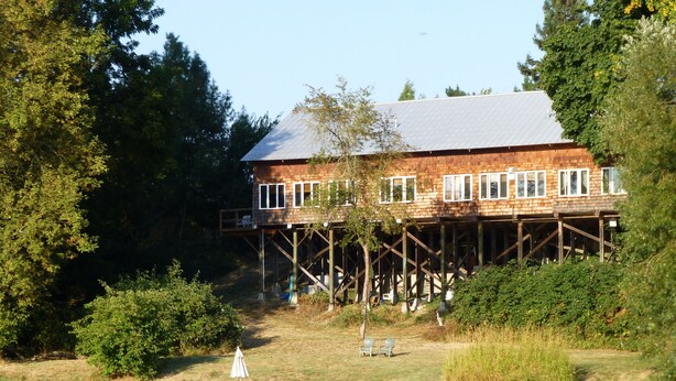 A wooden house with a deck, situated by a body of water, surrounded by trees.