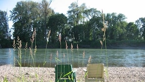 A riverside view with a chair and a bench facing the water, surrounded by tall grasses and trees.