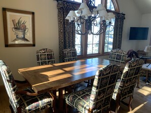 Comedor con mesa de madera, sillas estampadas, ventanas amplias y una lámpara de araña.