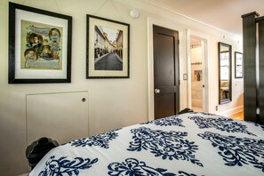A hotel room with a bed, framed pictures on the wall, a door leading to a bathroom, and a small trash bin.