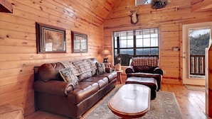 L’intérieur chaleureux d’un chalet avec un plafond en bois, un canapé en cuir, une table basse en bois et une grande fenêtre donnant sur le paysage extérieur.