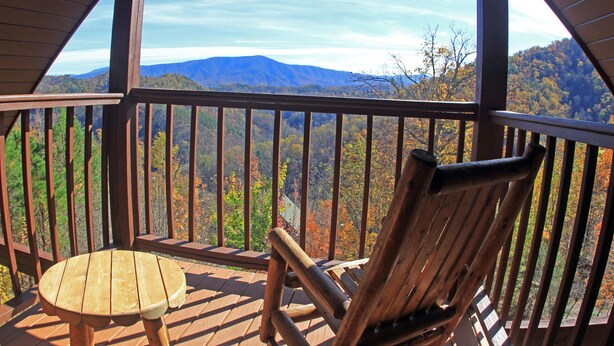 Une terrasse en bois avec une table et une chaise, offrant une vue sur un paysage montagneux aux couleurs automnales.