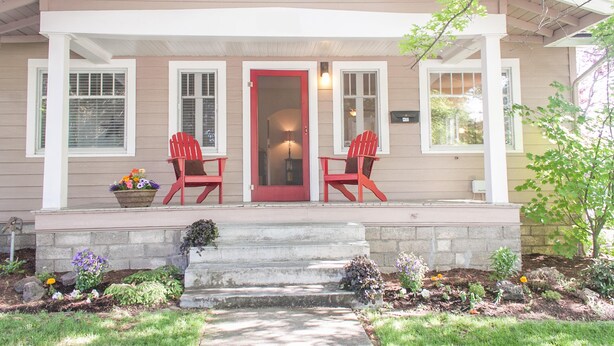 Una casa con un porche, dos sillas rojas, una puerta roja y un pequeño jardín con diversas plantas y flores.