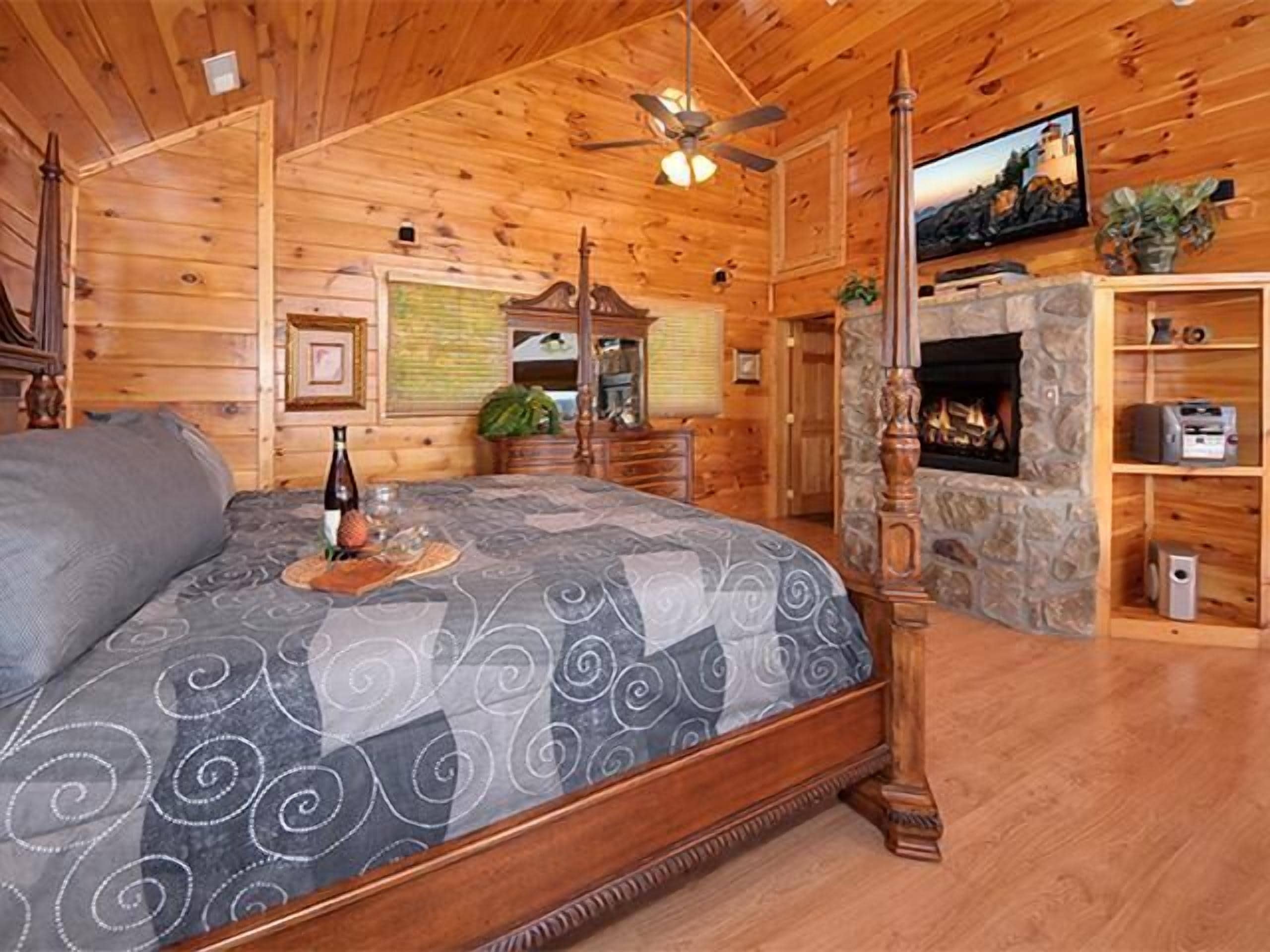 Un dormitorio tipo cabaña de madera con cama, chimenea, ventilador de techo y un revestimiento de piedra alrededor de la chimenea.