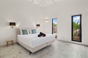 Un dormitorio con cama, dos luces fijadas a la pared, un taburete de madera pequeño y un amplio ventanal corredera que da a un espacio exterior.