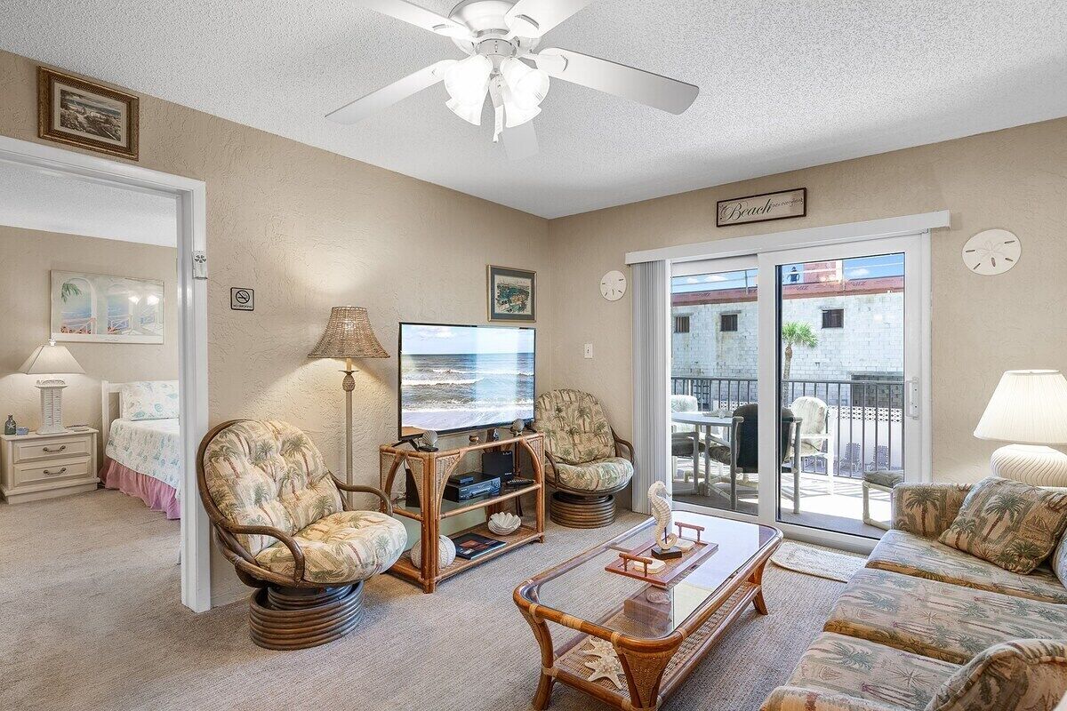Ein Wohnzimmer mit Deckenventilator, Fernseher, Sofa, Sessel und Blick ins Freie.