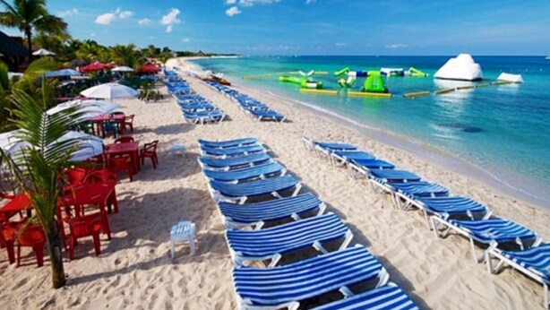 Una playa con filas de sillones reclinables a rayas azules y blancas y mesas rojas con sombrillas.