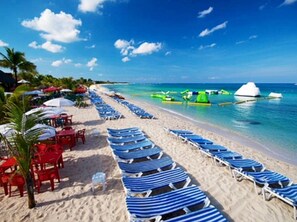 Una playa con filas de sillones reclinables a rayas azules y blancas y mesas rojas con sombrillas.