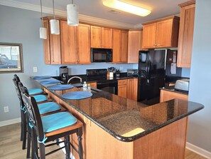 A modern kitchen with wooden cabinets, a black refrigerator, a microwave, and a built-in island with bar stools. - Beautiful condo. Short walk to the beach!  Linens & Towels included. Beds Made! (Ocean Isle Beach)