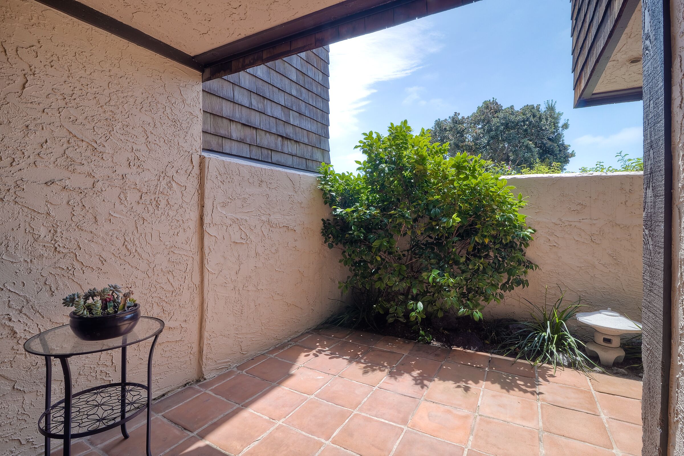 Un pequeño patio con suelo de baldosas, una mesa redonda con un cuenco de plantas y una planta en maceta contra una pared.