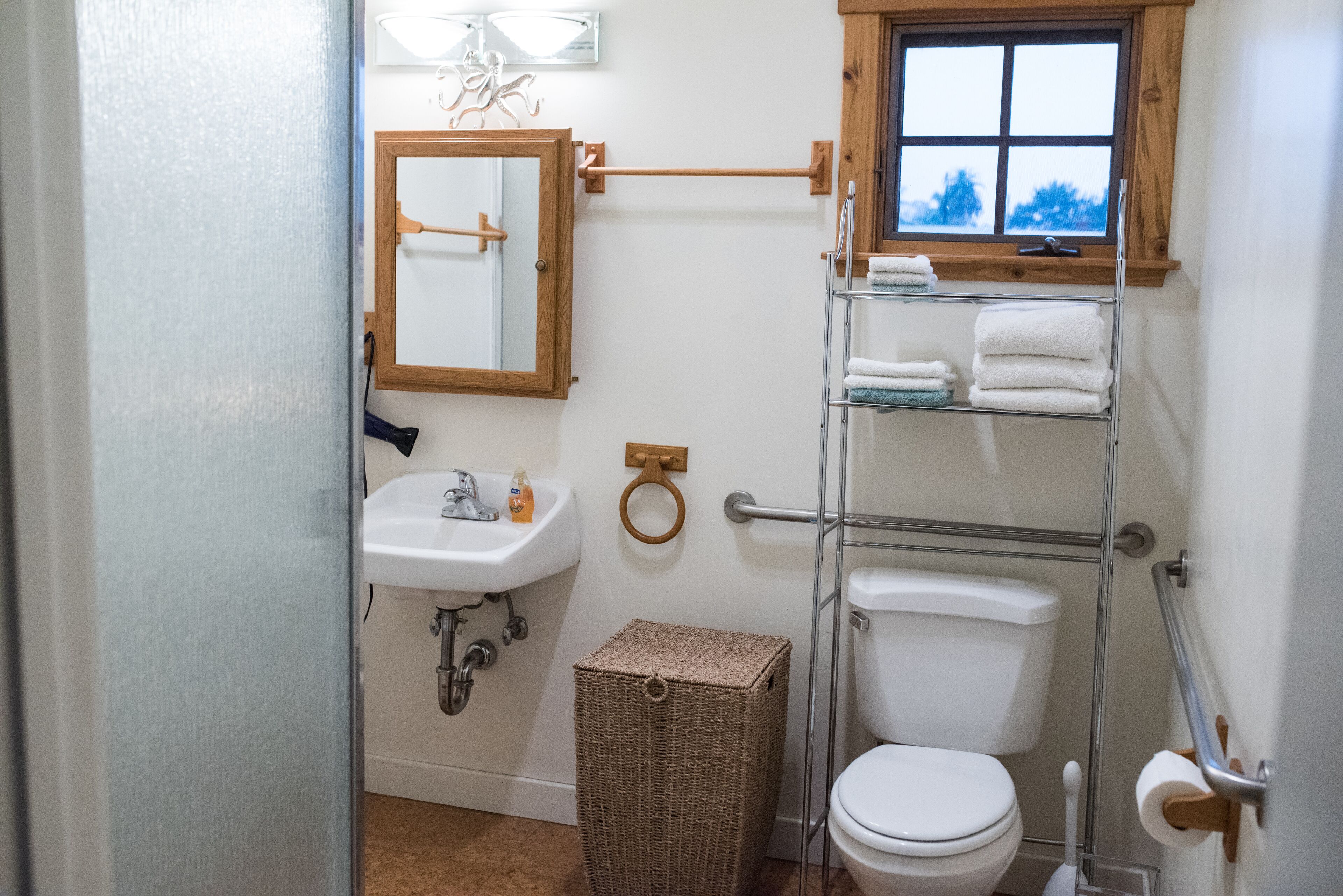 Une salle de bain avec un lavabo blanc, un miroir et des toilettes. Il y a une fenêtre au-dessus des toilettes, ainsi qu'un porte-serviettes avec des serviettes.