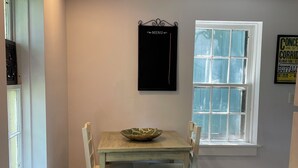 A dining area with a wooden table and white chairs, a black menu board, a window with blinds, and a sign on the wall. - The Moon and the Stars Cottage- in Town! (Nashville)