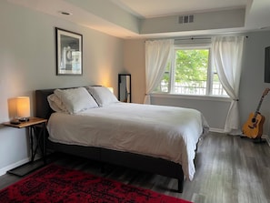 A bedroom with a bed, a nightstand with a lamp, a guitar, a window with curtains, and a red rug.