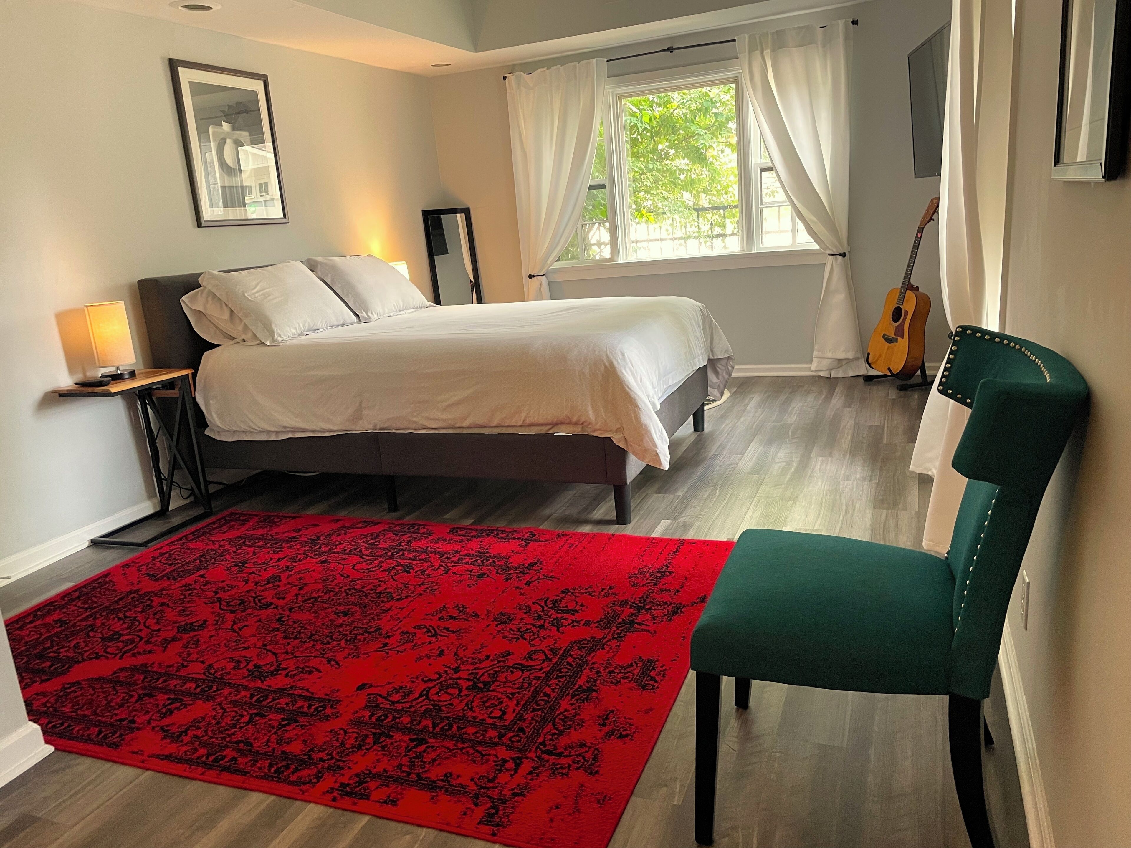 A bedroom with a bed, a red patterned rug, a green chair, a guitar, and a window with curtains.