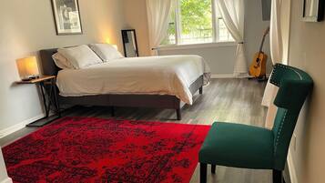 A bedroom with a bed, a red patterned rug, a green chair, a guitar, and a window with curtains.