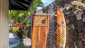 Une porte en bois, avec une planche de surf appuyée contre, un mur de pierre et un jardin tropical avec différentes plantes et un palmier.