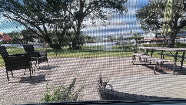 Une vue à travers une fenêtre munie d’un moustiquaire, donnant sur un parc avec des bancs, des arbres et une table de pique-nique.