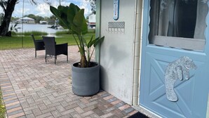 Un espace extérieur abrité, agrémenté d’une plante en pot, d’une porte ornée d’une figure en métal décorative, et offrant une vue sur un lac et des bâtiments en arrière-plan.