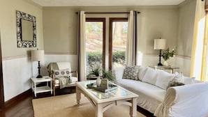 Un salón con sofá blanco, una mesa de centro de vidrio, una mesita auxiliar con lámpara y un espejo en la pared.