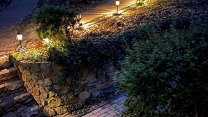 Un camino empedrado con muros de piedra y pequeñas luces, rodeado de árboles y vegetación.