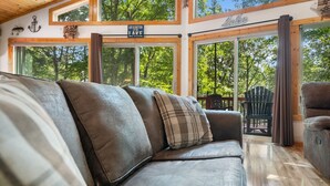 Un interior de cabaña acogedor con un amplio sofá de cuero, suelo de madera y grandes ventanas que dan a un bosque.