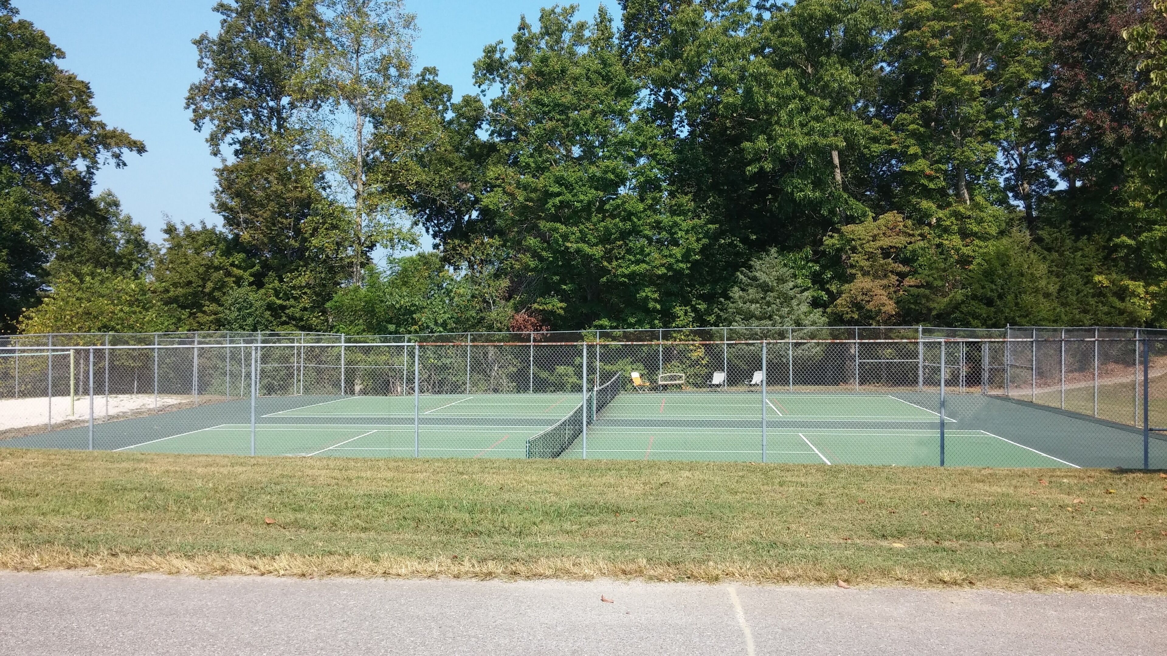 An outdoor tennis court with a chain-link fence, surrounded by trees and a grassy area.