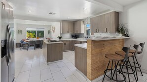 A modern kitchen with a central island, bar stools, and a dining area with a view of greenery. - The Compound: 2 Houses, 6 BR/4BA, Pool, Spa, Ping Pong, Pool Table, Events! (Indio)