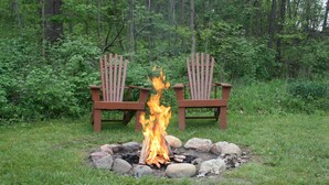 Two Adirondack chairs with a campfire between them in a grassy area surrounded by trees.