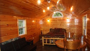 A cozy cabin interior with a wooden floor, a bed with a patterned bedspread, a dark-colored sofa, a wooden dining table with chairs, and a window with blinds.