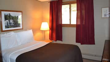 A hotel room with a bed, a nightstand with a lamp, a window with red curtains, and a framed picture on the wall.