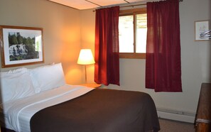 Une chambre d’hôtel avec un lit, une table de chevet avec une lampe, une fenêtre avec des rideaux rouges et un tableau encadré au mur.