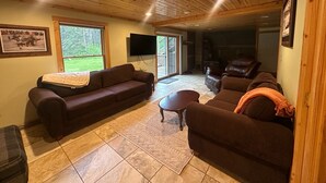 Un salon chaleureux avec un canapé marron, un fauteuil plus petit et une table basse. Un téléviseur est fixé au mur, et une fenêtre offre une vue sur la verdure à l’extérieur.