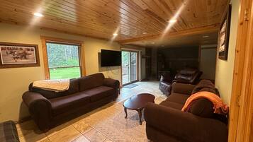 Un salon chaleureux avec un canapé marron, un fauteuil plus petit et une table basse. Un téléviseur est fixé au mur, et une fenêtre offre une vue sur la verdure à l’extérieur.