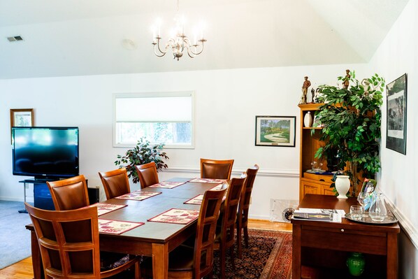 Zona pranzo con tavolo in legno, sedie, un lampadario, televisore e una finestra con luce naturale.