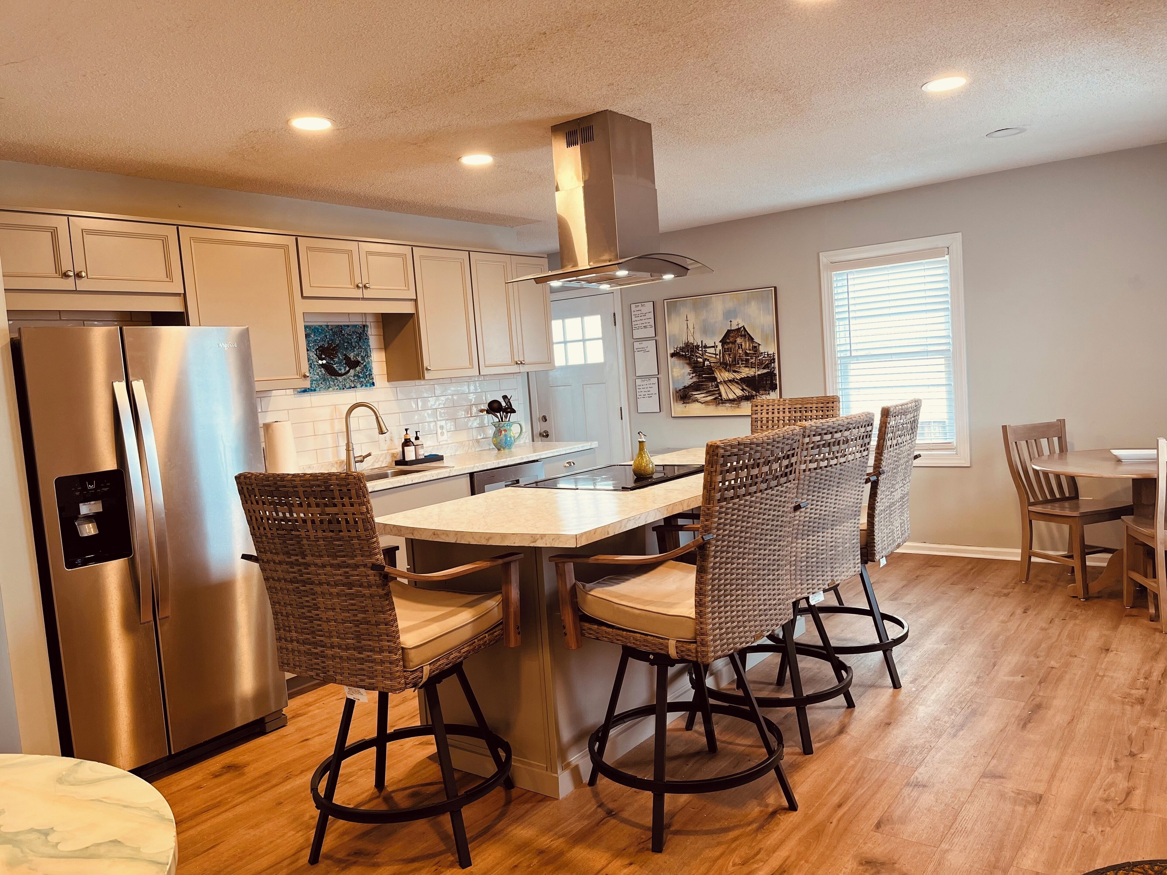 A modern kitchen with a stainless steel refrigerator, a central island with bar stools, and a dining area with wooden chairs.