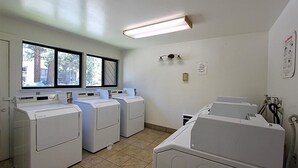 A laundry room with four front-loading washers and dryers, a large window with a view of trees and buildings, and a fluorescent light fixture. - Charming 2 Bedroom + Loft Condo Special Winter Ratesl (Mammoth Lakes)