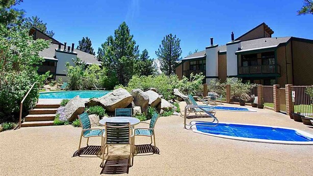 A pool area with lounge chairs and a table, surrounded by buildings and trees. - Charming 2 Bedroom + Loft Condo Special Winter Ratesl (Mammoth Lakes)