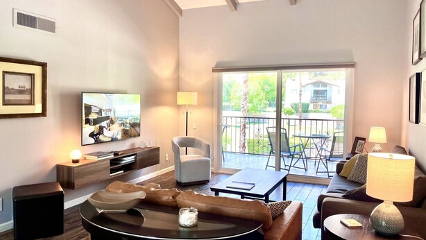 A modern living room with a flat-screen TV, comfortable seating, a balcony with outdoor furniture, and a large window providing a view of the outdoors. - Gorgeous condo with fabulous mountain and pool views in South Palm Springs (Palm Springs)