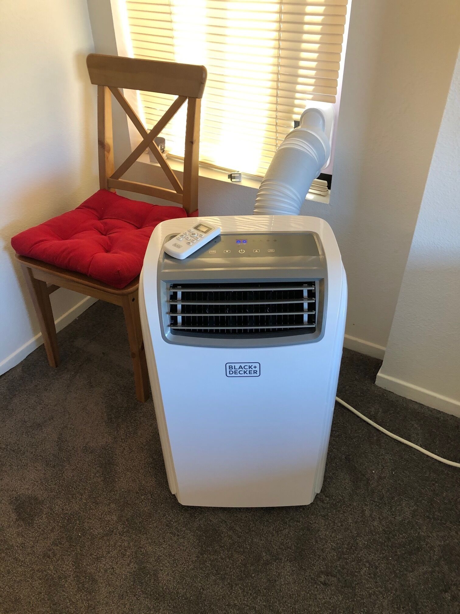 Un aire acondicionado portátil blanco de Black+Decker con control remoto, colocado al lado de una silla de madera con un cojín rojo, en una habitación con piso alfombrado y una ventana con persianas.