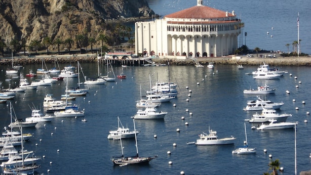 Un port de plaisance où sont amarrés de nombreux bateaux, un grand bâtiment avec un toit rouge et, au loin, une colline rocheuse.