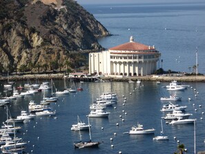 A marina with numerous boats docked, a large building with a red roof, and a rocky hillside in the background.