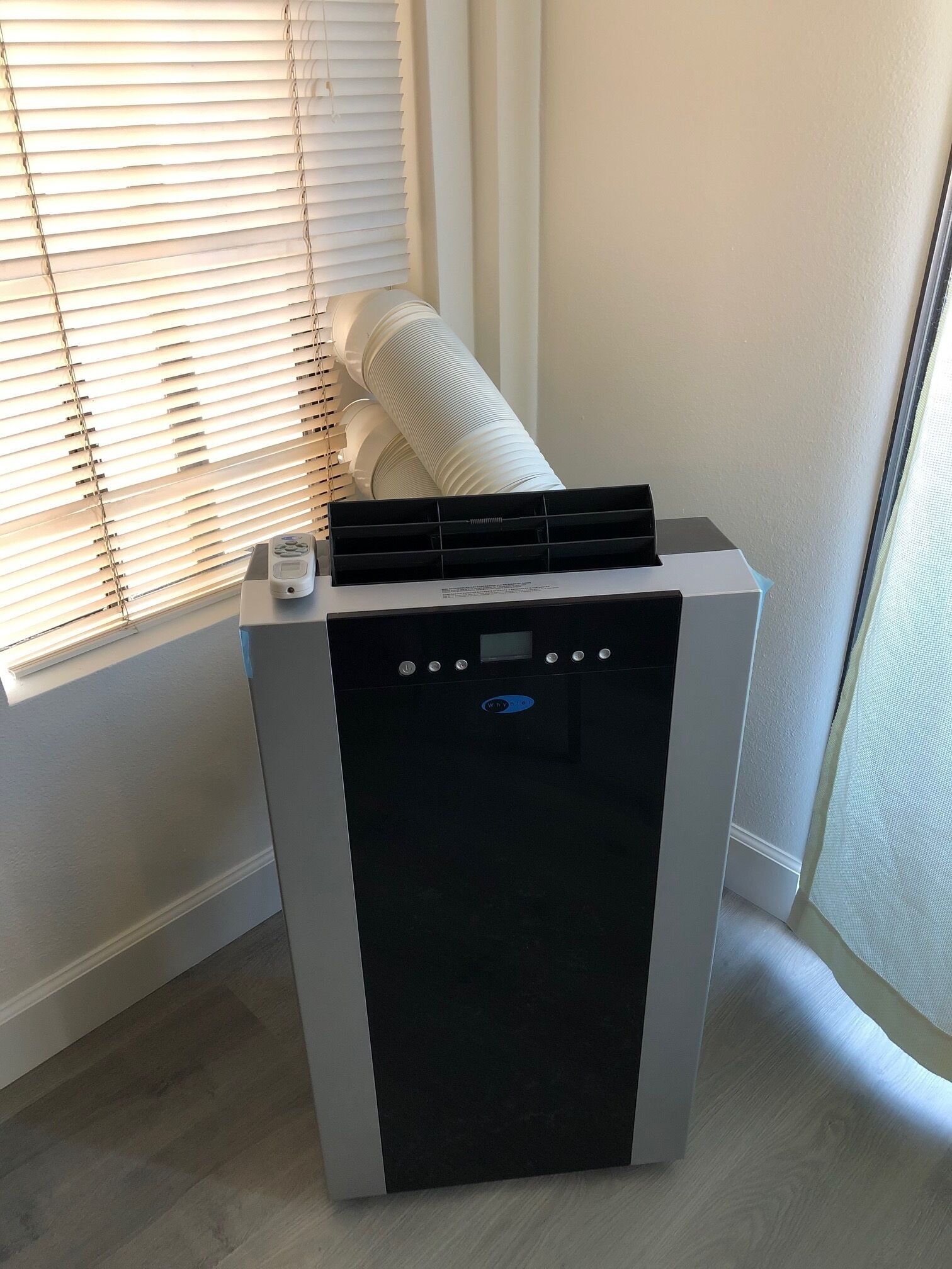 A compact, modern air purifier with a control panel and a filter compartment, placed near a window with blinds.