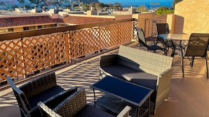 Une terrasse sur le toit, meublée de mobilier en osier, offrant une vue sur une ville perchée sur une colline et un ciel d’un bleu éclatant.