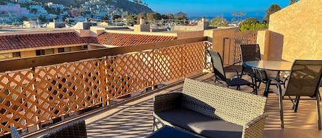 A rooftop terrace with wicker furniture, a view of a hillside town, and a clear blue sky.