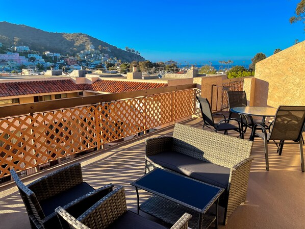A rooftop terrace with wicker furniture, a view of a hillside town, and a clear blue sky.